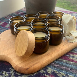 Pu&ograve; includere: Un tagliere di legno con pi&ugrave; piccoli barattoli di vetro ambrato pieni di una crema bianca. Un grande blocco di burro di karit&egrave; bianco &egrave; sul tagliere, insieme a pezzi pi&ugrave; piccoli di burro di karit&egrave;.