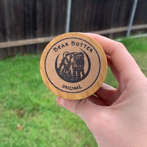 Pu&ograve; includere: Un contenitore rotondo in legno con un'etichetta di carta marrone che dice "Bear Butter Original" e presenta un'illustrazione in bianco e nero di un orso con una luna crescente dietro.