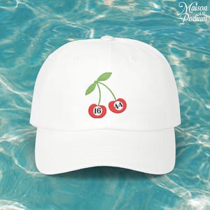 Puede incluir: Gorra de béisbol blanca con un diseño de cerezas bordadas con los números 16 y 44. La gorra tiene una visera curva y un logotipo de Maison de Podium.