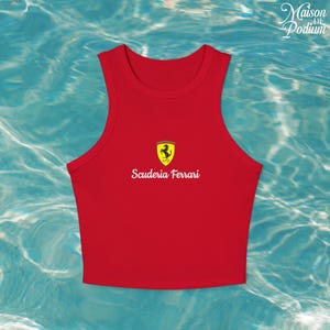 Puede incluir: Un top corto rojo con cuello redondo y el logo de Scuderia Ferrari. El logo presenta un escudo amarillo con un caballo negro y las palabras "Scuderia Ferrari" en blanco. El top es sin mangas.