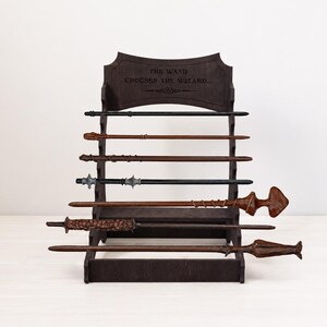 May include: A dark brown wooden display stand with five horizontal tiers holding seven distinct wands of varying lengths, colors, and textures. The top of the stand features the engraved text THE WAND CHOOSES THE WIZARD, with a decorative line element below.