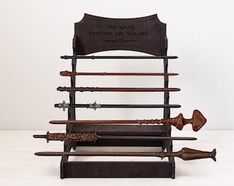 Puede incluir: Un expositor de madera marrón oscuro con cinco niveles horizontales que sostiene siete varitas distintas de diferentes longitudes, colores y texturas. La parte superior tiene el texto grabado THE WAND CHOOSES THE WIZARD con una línea decorativa.