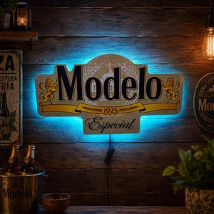 May include: A wall-mounted Modelo Especial sign featuring a tan background with black lettering, gold lion illustrations, and the text 1925. The sign is illuminated by a bright blue LED backlight, creating a glowing effect against the wooden wall.