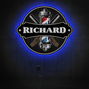 Puede incluir: Un letrero de neón azul con un poste de barbero y navajas cruzadas. El letrero tiene un fondo negro con el nombre "RICHARD" en texto blanco.