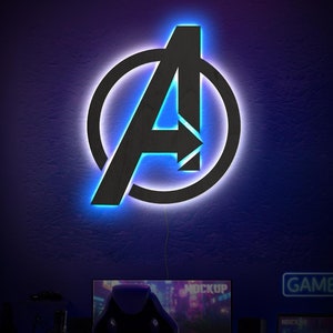 Könnte beinhalten: Ein an der Wand montiertes Avengers-Logo-Schild, bestehend aus einem schwarzen Buchstaben A in einem Kreis, beleuchtet durch eine hellblaue LED-Hintergrundbeleuchtung. Das Schild hängt über einem Computertisch in einem dunklen Raum.