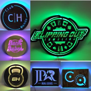Op de afbeelding: Vijf verlichte borden met verschillende ontwerpen. Het eerste bord is een zwarte cirkel met de letters "CH" in wit en de woorden "Club" en "Hendo" in wit. Het tweede bord is een zwarte cirkel met de woorden "Flipping Out Tumbling" in wit en een ontwerp van speelkaarten. Het derde bord is een zwarte cirkel met de woorden "Buck Rubbers" in wit. Het vierde bord is een zwarte cirkel met een witte kettlebell en de letters "EH" in zwart. Het vijfde bord is een zwarte rechthoek met de letters "JPR" in wit en de woorden "Real Estate" in wit.