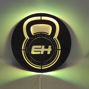 Op de afbeelding: Rond zwart houten bord met een uitgesneden ontwerp van een kettlebell. De kettlebell heeft de tekst "Elite Hall Fitness" in het ontwerp. Het bord wordt verlicht met een warm geel licht.