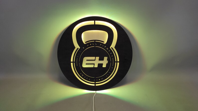 Op de afbeelding: Rond zwart houten bord met een uitgesneden ontwerp van een kettlebell. De kettlebell heeft de tekst "Elite Hall Fitness" in het ontwerp. Het bord wordt verlicht met een warm geel licht.