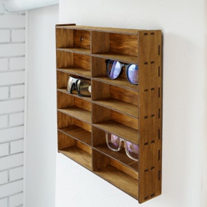 May include: A wooden wall-mounted shelf with multiple compartments for storing sunglasses. The shelf is made of dark brown wood and has a rustic look. There are two pairs of sunglasses on the shelf, one pair is black and the other is blue.