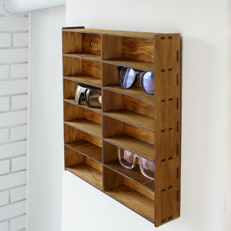 May include: A wooden wall-mounted shelf with multiple compartments for storing sunglasses. The shelf is made of dark brown wood and has a rustic look. There are two pairs of sunglasses on the shelf, one pair is black and the other is blue.