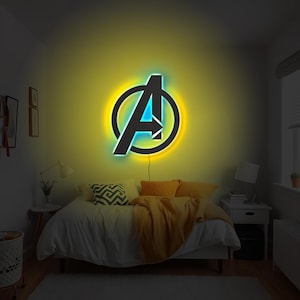 May include: A wall-mounted LED neon sign featuring the Avengers logo, characterised by a black letter A enclosed in a circle. The sign emits a vibrant yellow glow from behind the frame and a bright blue light from within the letter structure.