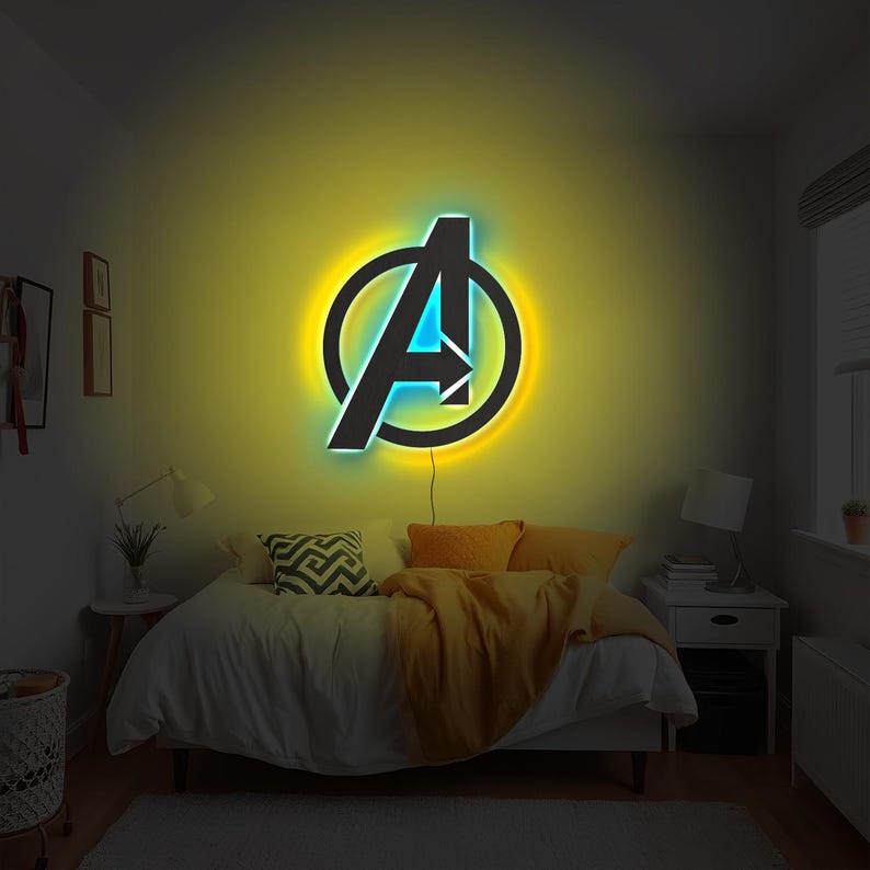 May include: A wall-mounted LED neon sign featuring the Avengers logo, characterised by a black letter A enclosed in a circle. The sign emits a vibrant yellow glow from behind the frame and a bright blue light from within the letter structure.