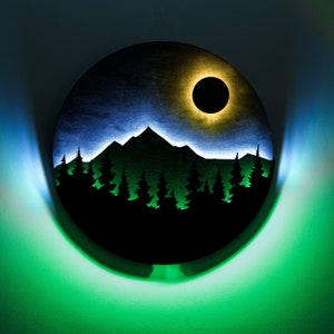 May include: A round wooden wall decoration featuring a black silhouette of a mountain range and trees against a blue and green sky. A yellow ring with a black circle in the centre is above the mountain range.