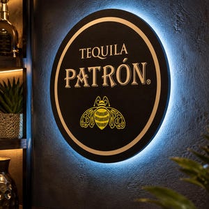 May include: A circular black wall sign featuring the white text TEQUILA PATRÓN and a yellow bee graphic. The sign is illuminated from behind with a bright blue LED glow, creating a halo effect against the dark, textured wall surface behind it.