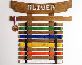 Op de afbeelding: Een donkerbruin houten displayrek voor vechtsportriemen met de naam OLIVER uitgesneden aan de bovenkant. Het rek heeft horizontale latten die worden vastgehouden door zwarte elastische banden, met groene, blauwe, gele, oranje en witte riemen.