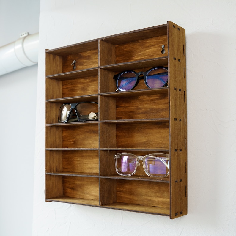 May include: A wooden wall-mounted shelf with multiple compartments for storing eyeglasses. The shelf is stained a dark brown color and has a decorative pattern on the side. There are three pairs of eyeglasses displayed on the shelf.