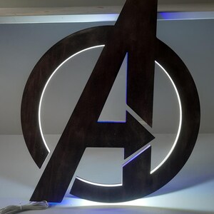 May include: A dark brown wooden Avengers logo wall sign featuring a circular frame and a central letter A. The sign is illuminated from behind with a bright white light that creates a glowing halo effect around the edges of the wooden cutout.