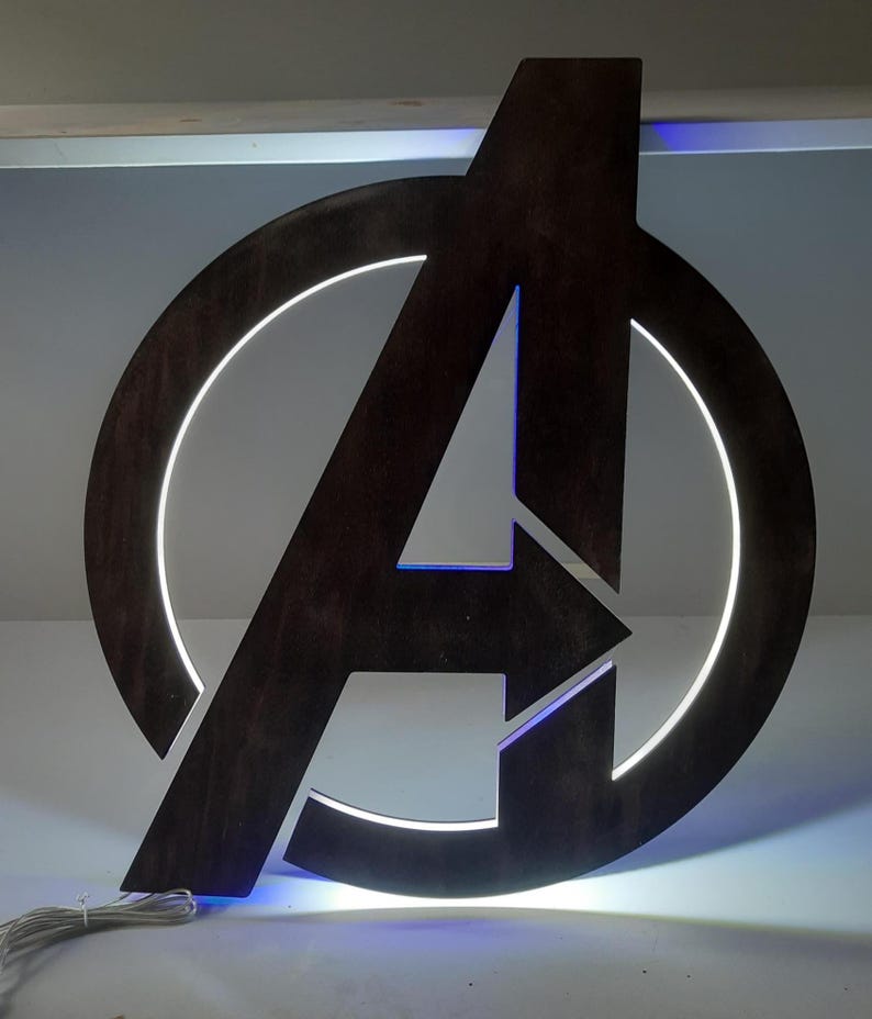 May include: A dark brown wooden Avengers logo wall sign featuring a circular frame and a central letter A. The sign is illuminated from behind with a bright white light that creates a glowing halo effect around the edges of the wooden cutout.