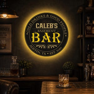 Może przedstawiać: Okrągły podświetlany znak ścienny z napisem CALEB'S BASEMENT BAR, COLD DRINKS & GOOD PEOPLE, AUSTIN, TX oraz EST. 2010. Znak posiada czarne tło z żółtymi literami oraz ozdobnymi elementami graficznymi.