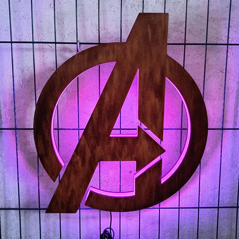May include: A dark brown wooden wall sign featuring the Avengers logo, which consists of a capital letter A enclosed within a circular frame. The sign is illuminated from behind with a vibrant purple LED light that glows around the edges.