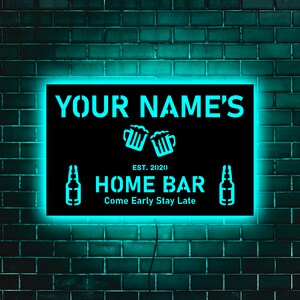 May include: A black neon sign with white text that reads "YOUR NAME'S HOME BAR EST. 2020 Come Early Stay Late". The sign is illuminated with a bright turquoise light and features a graphic of two beer mugs and two beer bottles.