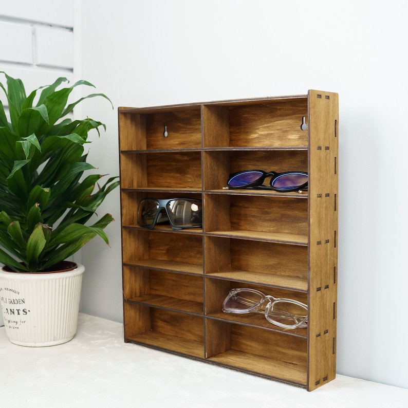 May include: A wooden wall-mounted shelf with eight compartments for storing sunglasses. The shelf is made of light brown wood and has a rustic finish.