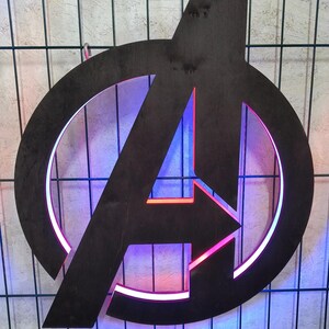 May include: A dark brown wooden Avengers logo wall sign featuring a circular frame and a central letter A. The cutout design is illuminated from behind with vibrant red and blue LED lighting, creating a glowing effect around the edges of the wood.