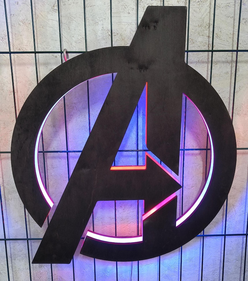 May include: A dark brown wooden Avengers logo wall sign featuring a circular frame and a central letter A. The cutout design is illuminated from behind with vibrant red and blue LED lighting, creating a glowing effect around the edges of the wood.