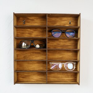 May include: A wooden wall-mounted shelf with nine compartments for storing eyeglasses. Three pairs of eyeglasses are displayed on the shelf, including a pair of black sunglasses, a pair of blue-tinted glasses, and a pair of clear glasses.