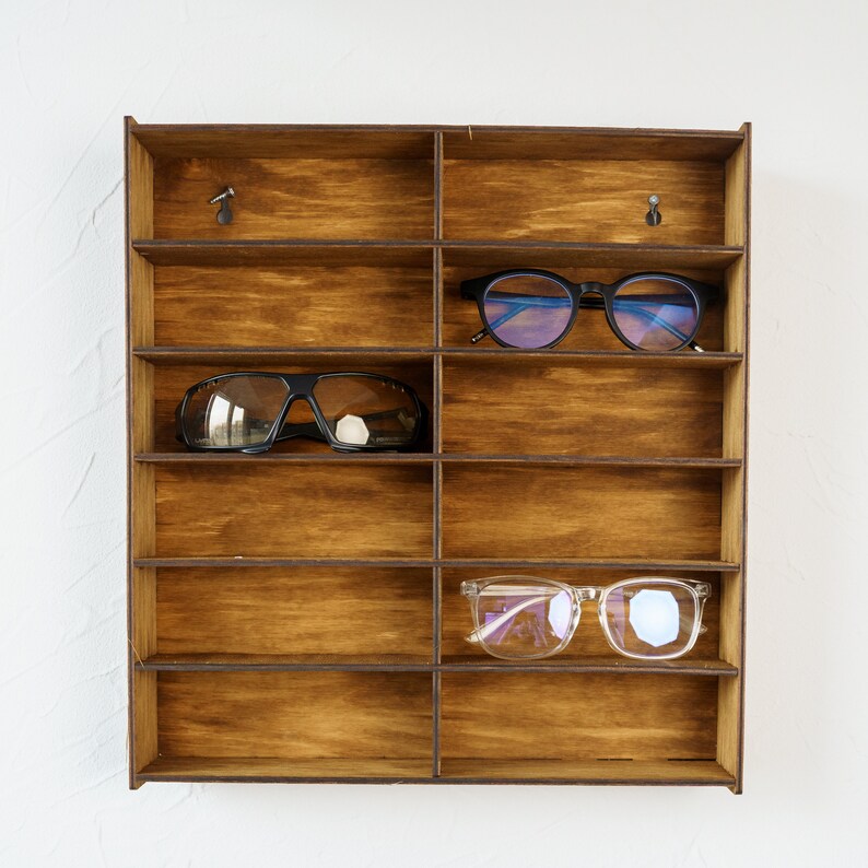 May include: A wooden wall-mounted shelf with nine compartments for storing eyeglasses. Three pairs of eyeglasses are displayed on the shelf, including a pair of black sunglasses, a pair of blue-tinted glasses, and a pair of clear glasses.