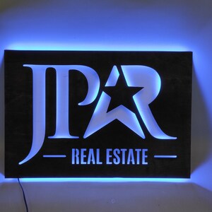 Op de afbeelding: Een zwart metalen bord met een witte uitsparing van de letters "JPAR" en een ster. Het bord is verlicht met blauwe LED-verlichting en heeft de tekst "REAL ESTATE" onder het logo.