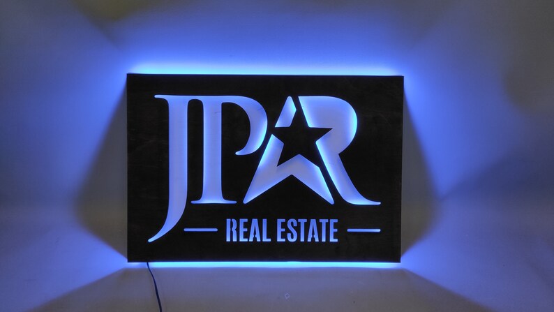 Op de afbeelding: Een zwart metalen bord met een witte uitsparing van de letters "JPAR" en een ster. Het bord is verlicht met blauwe LED-verlichting en heeft de tekst "REAL ESTATE" onder het logo.