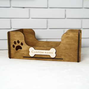 Peut inclure: Un lit pour animaux de compagnie en bois brun avec une empreinte de patte et une &eacute;tiquette en forme d'os avec l'inscription "CUSTOM NAME". Le lit a un profil bas.