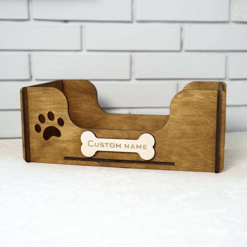 Peut inclure: Un lit pour animaux de compagnie en bois brun avec une empreinte de patte et une &eacute;tiquette en forme d'os avec l'inscription "CUSTOM NAME". Le lit a un profil bas.