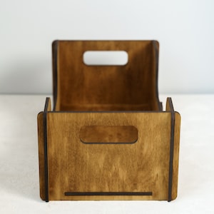 Peut inclure: Une caisse de rangement en bois avec une finition marron fonc&eacute;. La caisse a une forme rectangulaire avec un dos incurv&eacute; et deux d&eacute;coupes rectangulaires pour les poign&eacute;es &agrave; l'avant. Le grain du bois est visible, et la caisse semble vide.