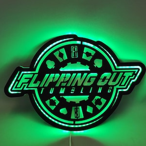 Op de afbeelding: Een zwart en groen verlicht bord met de tekst "Flipping Out Tumbling" in wit. Het bord heeft een cirkelvormig ontwerp met speelkaartsymbolen en een tandwiel in het midden.