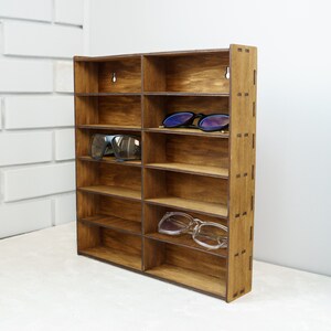 May include: A wooden wall-mounted shelf with 10 compartments for storing sunglasses. The shelf is made of dark brown wood and has a natural wood grain finish. There are two pairs of sunglasses on the shelf.