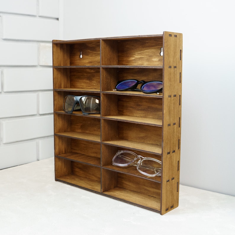 May include: A wooden wall-mounted shelf with 10 compartments for storing sunglasses. The shelf is made of dark brown wood and has a natural wood grain finish. There are two pairs of sunglasses on the shelf.