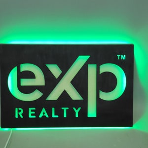Op de afbeelding: Een zwart bord met een witte uitsparing van het woord "exp" en de woorden "REALTY" eronder. Het bord is verlicht met groene LED-verlichting rond de omtrek.