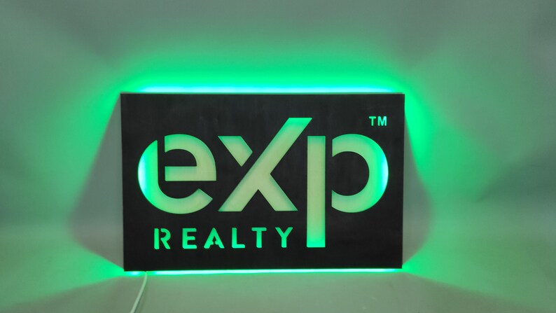 Op de afbeelding: Een zwart bord met een witte uitsparing van het woord "exp" en de woorden "REALTY" eronder. Het bord is verlicht met groene LED-verlichting rond de omtrek.