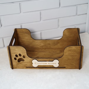 Peut inclure: Un panier pour animaux de compagnie en bois avec une d&eacute;coupe en forme de patte et une &eacute;tiquette en forme d'os avec l'inscription "CUSTOM NAME". Le lit a une base rectangulaire avec des c&ocirc;t&eacute;s incurv&eacute;s et des poign&eacute;es.
