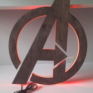 May include: A dark grey wooden Avengers logo wall sign featuring a circular frame and a central letter A. The sign is illuminated from behind with a vibrant red LED light glow. A black power cord with a USB connector extends from the bottom edge.