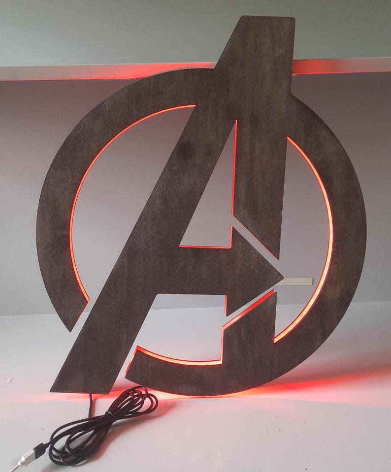 May include: A dark grey wooden Avengers logo wall sign featuring a circular frame and a central letter A. The sign is illuminated from behind with a vibrant red LED light glow. A black power cord with a USB connector extends from the bottom edge.