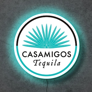May include: A circular wall sign featuring the Casamigos Tequila logo with a teal agave plant graphic centered above the black text CASAMIGOS and the word Tequila in a serif font, all surrounded by a glowing teal light border on a dark background.