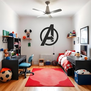 May include: A large black Avengers logo wall sign mounted on a white wall in a bedroom. The sign features the iconic letter A enclosed within a circle. The room includes a black desk, a blue office chair, a bed with red bedding, and a red area rug.