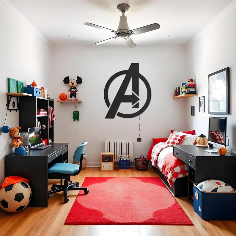 May include: A large black Avengers logo wall sign mounted on a white wall in a bedroom. The sign features the iconic letter A enclosed within a circle. The room includes a black desk, a blue office chair, a bed with red bedding, and a red area rug.