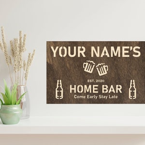 May include: A brown wooden sign with white text that reads "YOUR NAME'S HOME BAR EST. 2020 Come Early Stay Late". The sign features a graphic of two beer mugs clinking and two beer bottles.