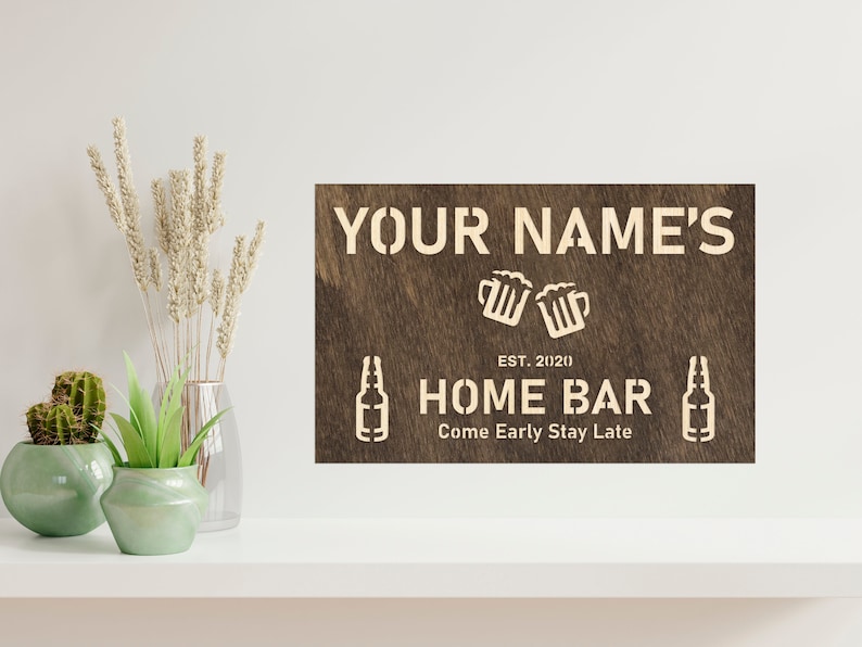 May include: A brown wooden sign with white text that reads "YOUR NAME'S HOME BAR EST. 2020 Come Early Stay Late". The sign features a graphic of two beer mugs clinking and two beer bottles.