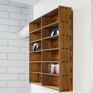 May include: A wooden wall-mounted shelf with multiple compartments for storing glasses or sunglasses. The shelf is stained a dark brown color and has a decorative pattern on the side.