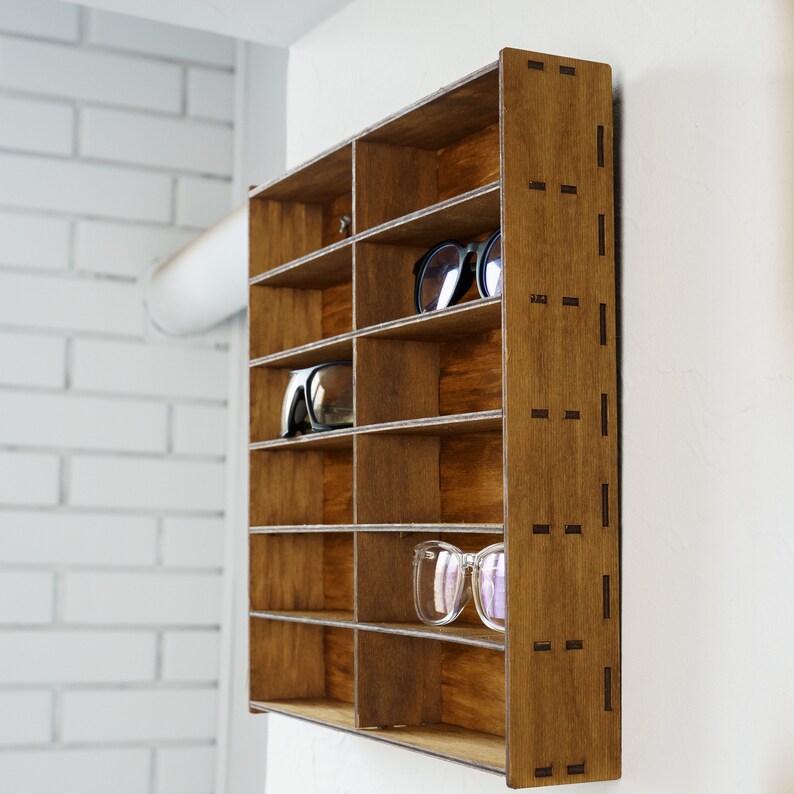 May include: A wooden wall-mounted shelf with multiple compartments for storing glasses or sunglasses. The shelf is stained a dark brown color and has a decorative pattern on the side.